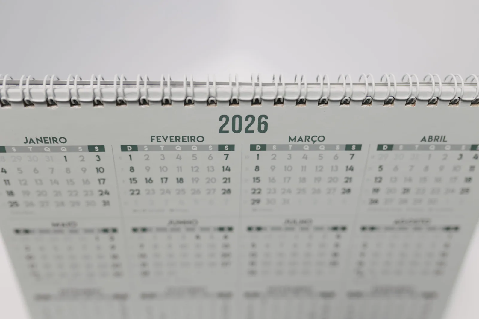 Close-up of a 2026 spiral-bound desk calendar with months in Portuguese. Days between dates holidays: past and future windows