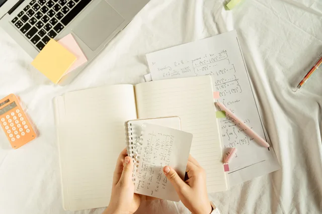 Hands holding a notebook with math equations, calculator, and laptop on a study desk. Spreadsheet and API tools formula testing for parity